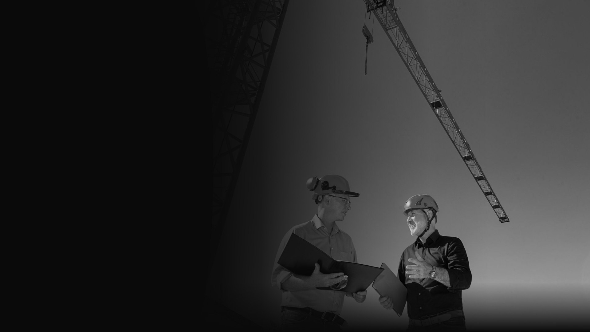Zwei Männer mit Schutzhelmen im Gespräch auf einer Baustelle, im Hintergrund ein Kranausleger